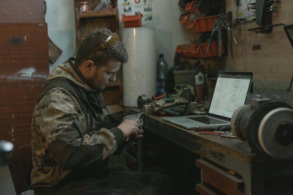 A man intensely focuses on repairing a part at his industrial workspace beside a laptop.